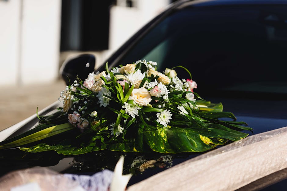 A floral arrangement featuring a bouquet of fresh cream and white roses, delicate white daisies, and small baby's breath, all accented with lush green foliage and long, broad leaves. The flowers are a