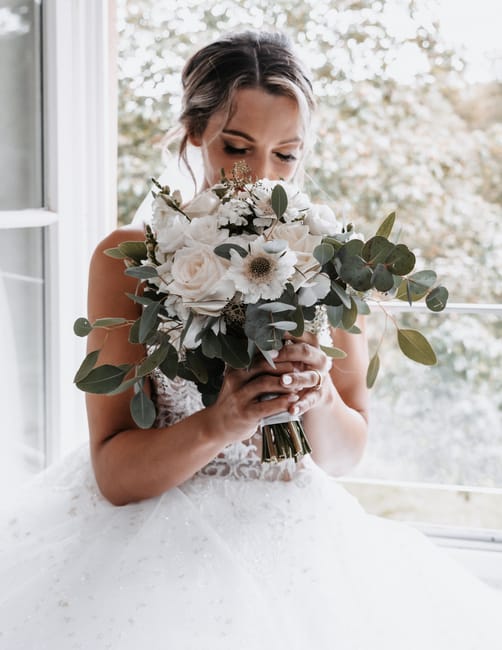 A bride holding a large floral bouquet composed of white roses, anemones with black centers, and eucalyptus leaves. The bouquet is wrapped with a dark ribbon at the stems. She is wearing a lace weddin
