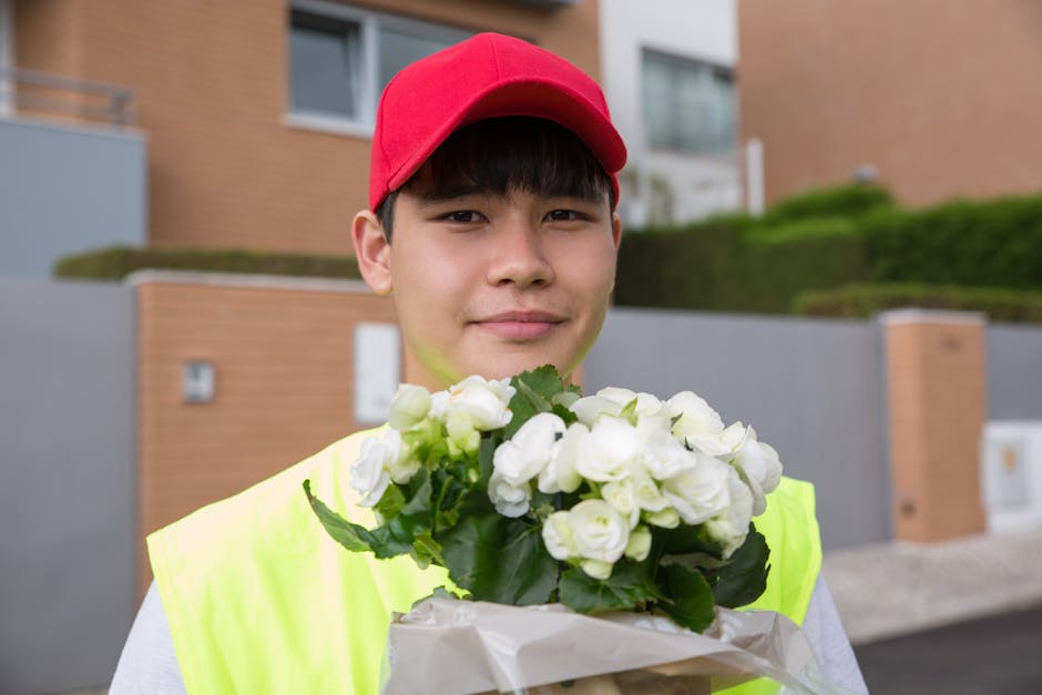A young person wearing a red cap and a high-visibility yellow vest holds a bouquet of fresh white roses wrapped in brown craft paper. The flowers have lush green leaves, and the petals appear soft and