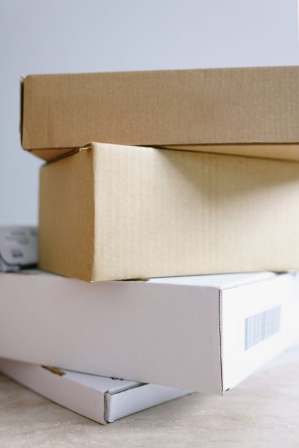 The image shows a close-up of three cardboard boxes stacked on top of each other, with the top box being plain brown, the middle one a slightly lighter beige, and the bottom one white with a barcode. 