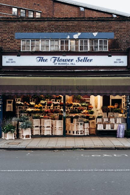 The exterior of The Flower Seller shop on Muswell Hill features a dark awning with the shop's name prominently displayed in elegant script, flanked by the phrase